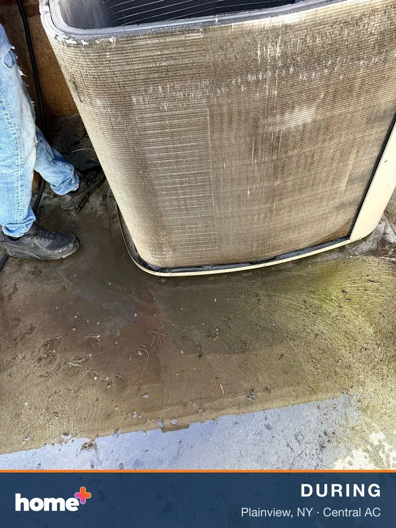 A technician's lower body is seen next to an outdoor AC unit, which is actively being cleaned with water, causing puddles on the ground.